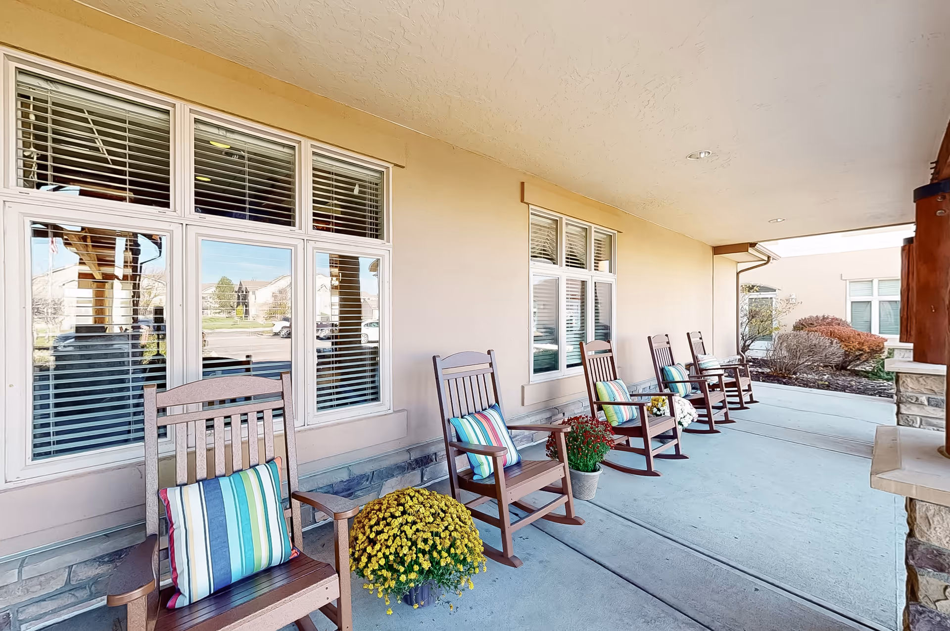 Covered outdoor patio area at MeadowView of Greeley with a row of wooden rocking chairs, each with a colorful striped cushion. Potted flowers are placed between some of the chairs. The patio is adjacent to a beige building with multiple windows and overlooks a landscaped area with bushes and a parking lot in the background.
