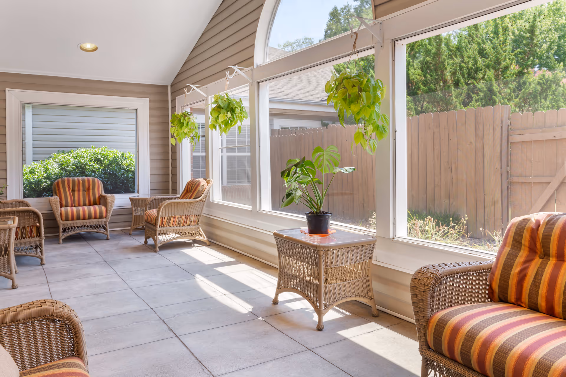 A bright sunroom with large windows and a glass door looking out to a fenced garden. The room features wicker furniture with striped orange and brown cushions, a wicker side table with a potted plant, and hanging green plants near the windows.