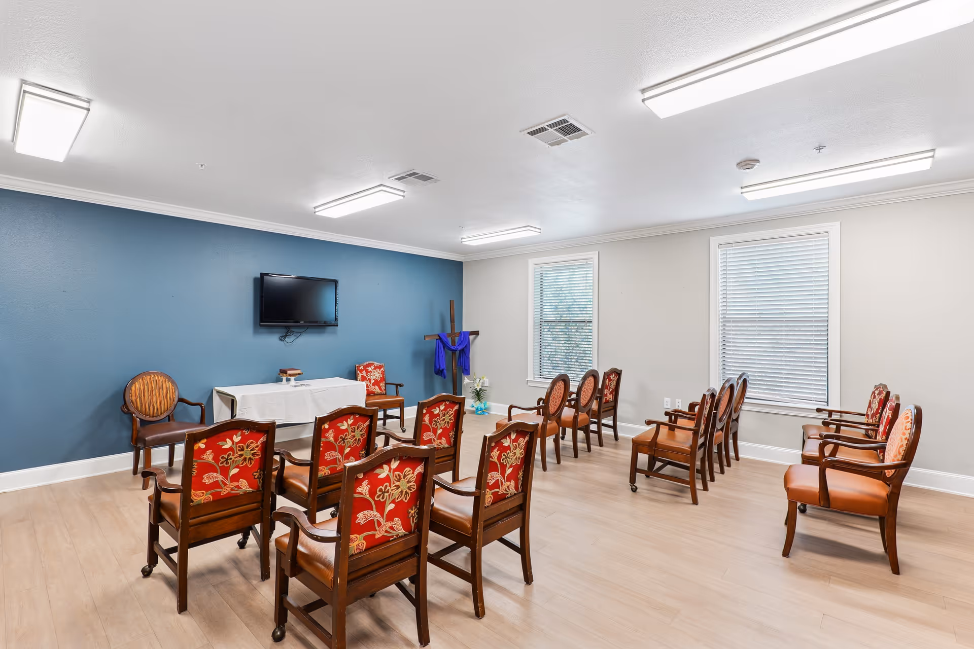 A small chapel or meeting room with wooden chairs arranged in rows facing a table covered with a white cloth. A flat-screen TV is mounted on a blue accent wall behind the table, and a wooden cross draped with a purple cloth stands in the corner near two windows with blinds. The room has light-colored wood flooring and bright overhead lighting.