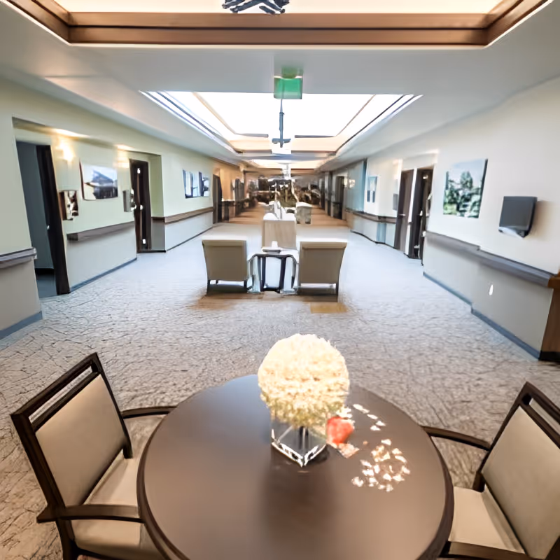 A spacious, well-lit hallway in a senior living facility with a round table and two chairs in the foreground. The table has a decorative white flower arrangement. The hallway features carpeted floors, wall-mounted handrails, framed pictures on the walls, and seating areas with armchairs further down the corridor. Skylights provide natural light from above.