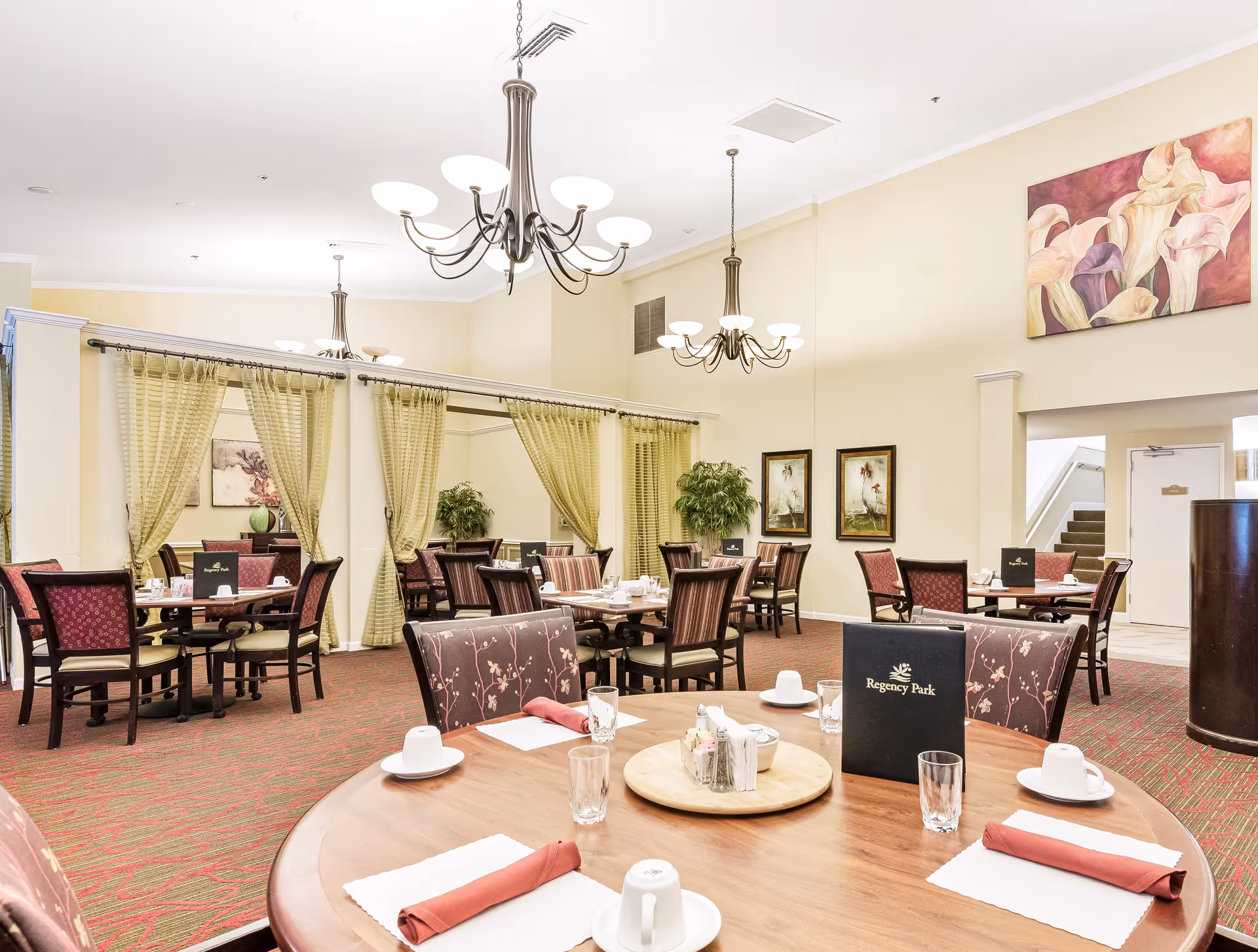 A bright and spacious dining room in Regency Park facility with round wooden tables set with white cups, glasses, napkins, and menus. The room features red patterned carpet, upholstered chairs, hanging chandeliers, green curtains, potted plants, and framed artwork on cream-colored walls.
