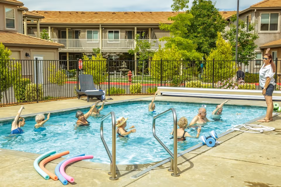A group of elderly people participating in a water exercise class in an outdoor swimming pool, led by an instructor standing at the poolside. The pool is surrounded by a fence, with residential buildings and greenery in the background. Pool noodles and water weights are visible on the pool deck.