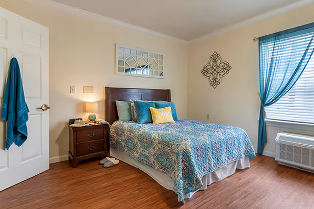 Bright bedroom with a double bed dressed in patterned teal bedding, a wooden nightstand with a lamp, wall decor, and a window with teal curtains.