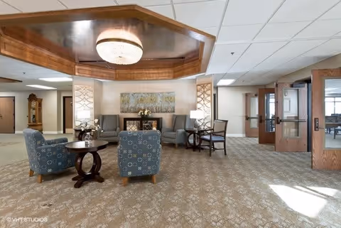 A spacious and well-lit common area with patterned carpet, several upholstered chairs arranged around small wooden tables, a decorative ceiling with a chandelier, and a large painting on the far wall. There are wooden doors on the right side leading to another room.