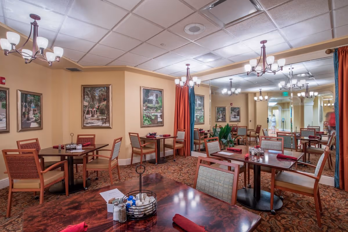A cozy dining room in Luxe Senior Living at Wellington featuring multiple wooden tables with chairs arranged around them. Each table has red cloth napkins, salt and pepper shakers, and a centerpiece. The room has warm beige walls adorned with framed artwork, a patterned carpet, and ceiling light fixtures. A large mirror covers one wall, reflecting the room and adding depth.