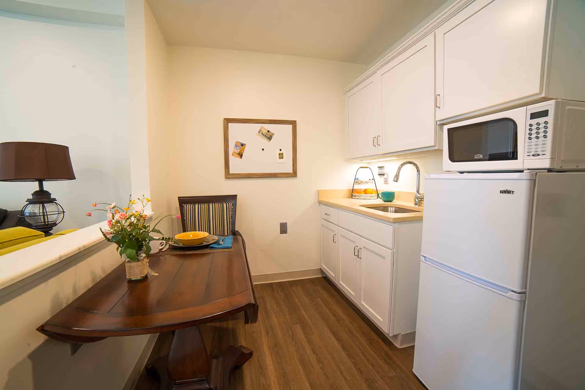 A small kitchen area with white cabinets, a countertop with a sink, a white microwave on top of a white refrigerator, and a wooden table set against a half wall. The table has a vase with flowers, a yellow bowl, a cup, and a placemat with a napkin and utensils. A chair with a striped cushion is placed at the table, and a bulletin board with a few pinned items hangs on the wall above the table.