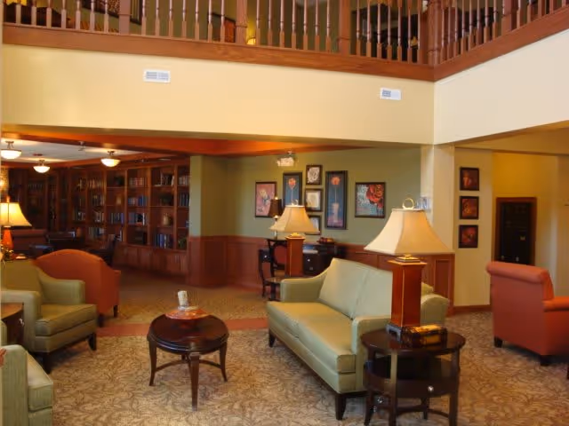 A spacious living room area in a senior living facility with comfortable seating including green sofas and armchairs, wooden side tables with lamps, framed artwork on the walls, and a built-in wooden bookshelf filled with books. The room has a high ceiling with a wooden balcony railing above and warm lighting.