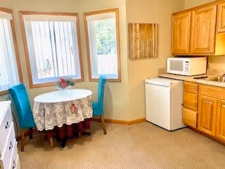 A small dining area with a round table covered with a white lace tablecloth and two teal upholstered chairs. The area is next to two windows with vertical blinds, allowing natural light to enter. To the right, there is a kitchenette with wooden cabinets, a microwave, and a small refrigerator. The walls are painted a light beige color.