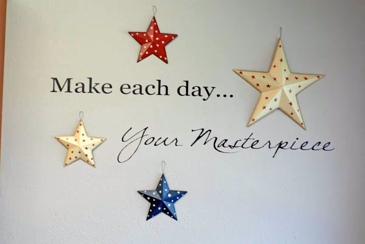 A wall decoration featuring five hanging stars in red, white, and blue colors with small star patterns, surrounding the inspirational quote 'Make each day... Your Masterpiece' written in black text on a light-colored wall.