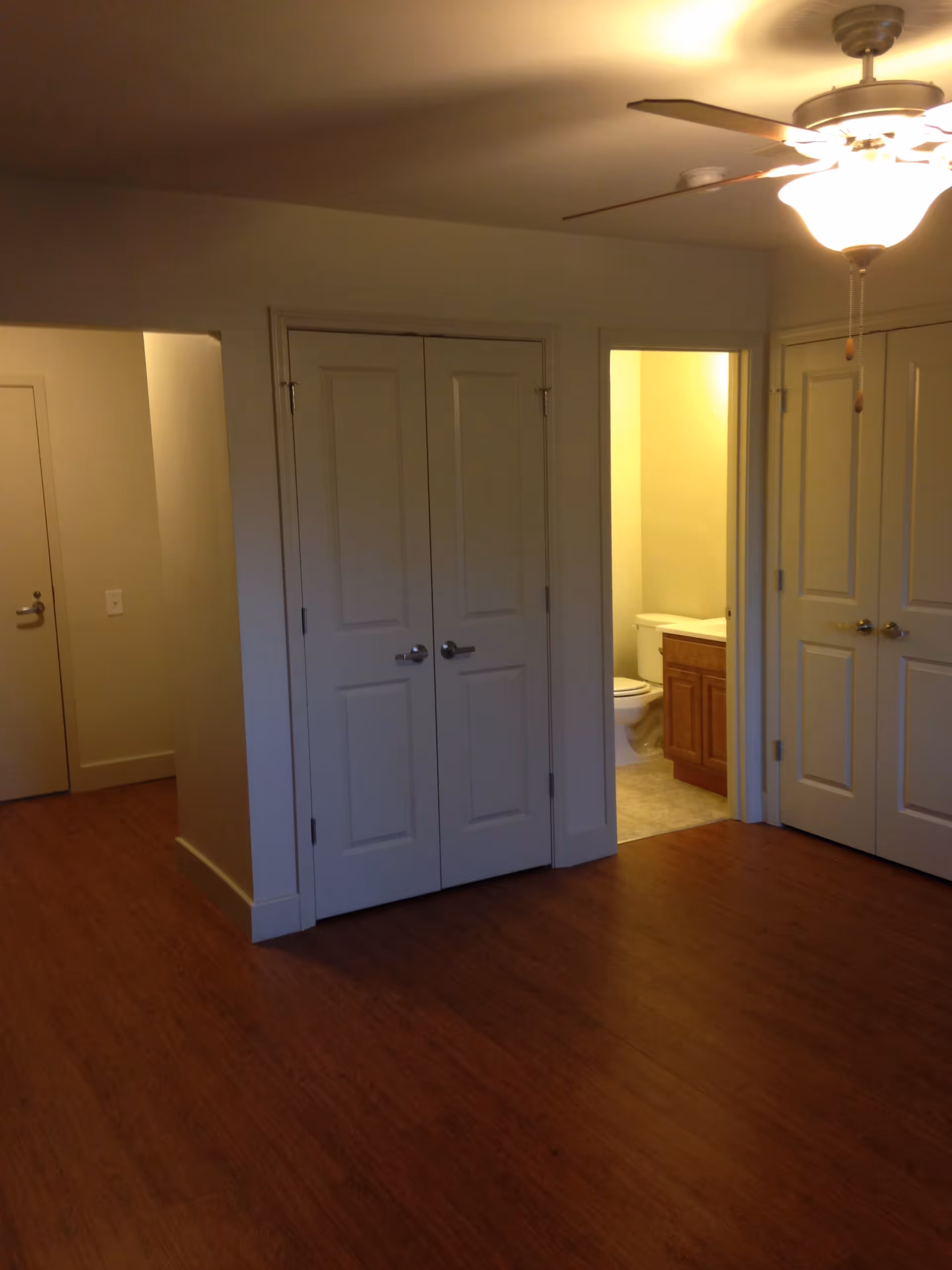 Interior view of a room with wooden flooring, white walls, and multiple closed white doors. One door is open, revealing a bathroom with a toilet and a wooden vanity. A ceiling fan with a light fixture is visible on the ceiling.