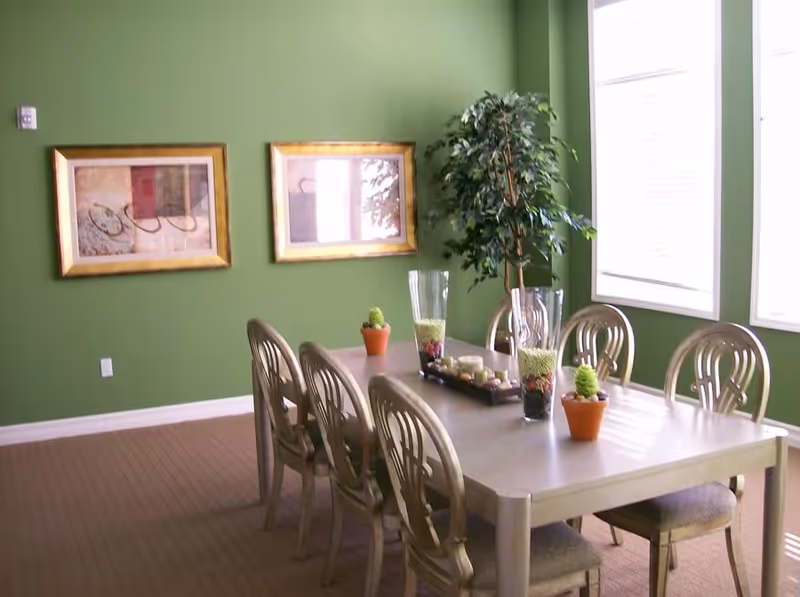 A dining room with a long rectangular table surrounded by six chairs. The table is decorated with small potted plants and tall glass vases filled with decorative items. The walls are painted green and have two framed abstract paintings. Large windows on the right side let in natural light.