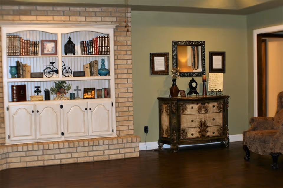 Living room with a built-in white bookcase set into a brick wall, an ornate chest with mirror and decor, and an upholstered chair on wood flooring.