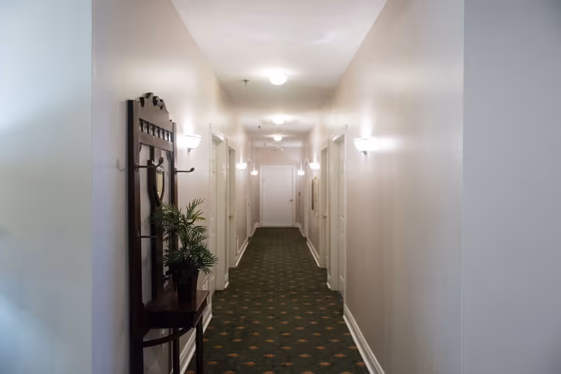 A long, narrow hallway in a retirement community with beige walls, green carpet with a subtle pattern, and several white doors on both sides. The hallway is lit by ceiling lights and wall sconces. A wooden hall tree with a mirror and a potted plant is positioned on the left side near the foreground.