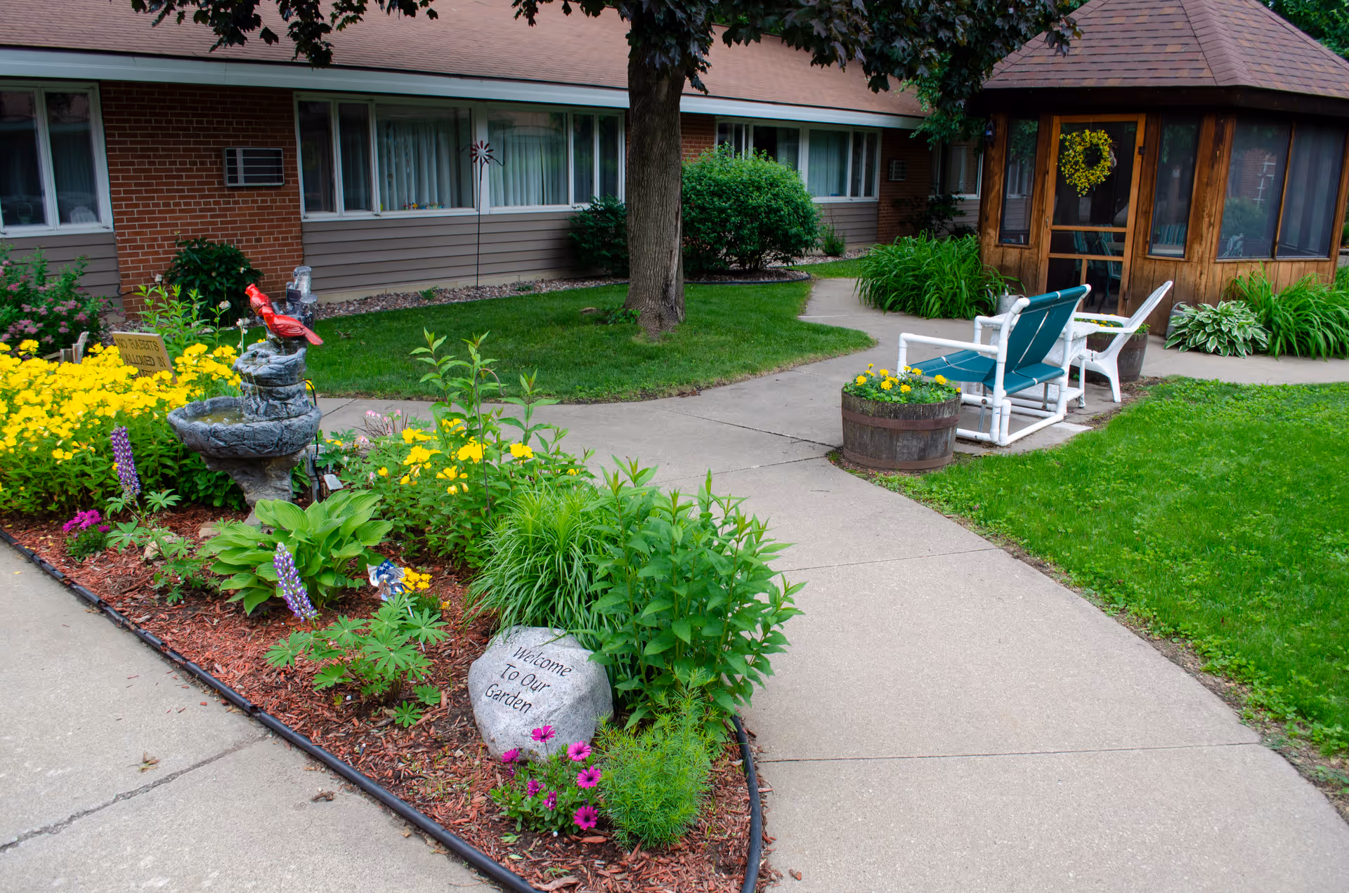A well-maintained garden area with colorful flowers, a small stone fountain with a red bird decoration, and a rock engraved with 'Welcome To Our Garden'. There is a paved walkway leading to a wooden gazebo with a wreath on the door, and two chairs with a small table nearby. The background shows a brick and siding building with windows and greenery.