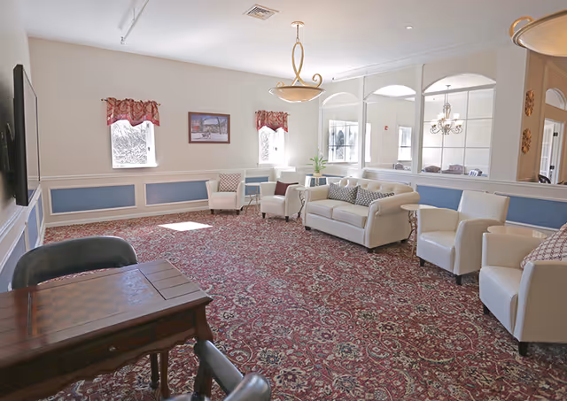 A spacious living room with a patterned red carpet, white walls with blue paneling, and multiple white armchairs and a white sofa arranged around the room. There is a wooden table with a checkerboard pattern in the foreground, two small windows with red valances, a framed picture on the wall, and a large mirror reflecting a chandelier and dining area. The room is well-lit with natural light and ceiling lights.