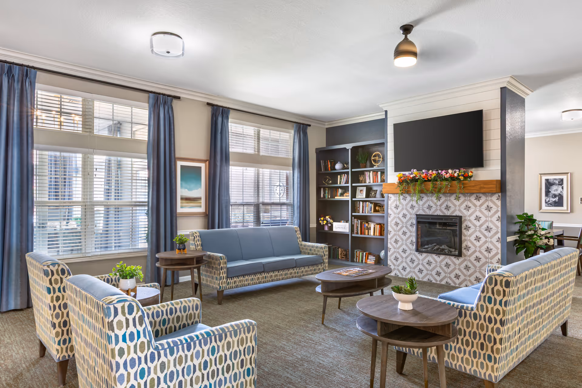 A bright and cozy living room with large windows covered by blue curtains, patterned armchairs, and a blue sofa arranged around two wooden coffee tables. There is a fireplace with decorative tiles and a flat-screen TV mounted above it. A bookshelf filled with books and decorative items is next to the fireplace, and a few potted plants add greenery to the space.