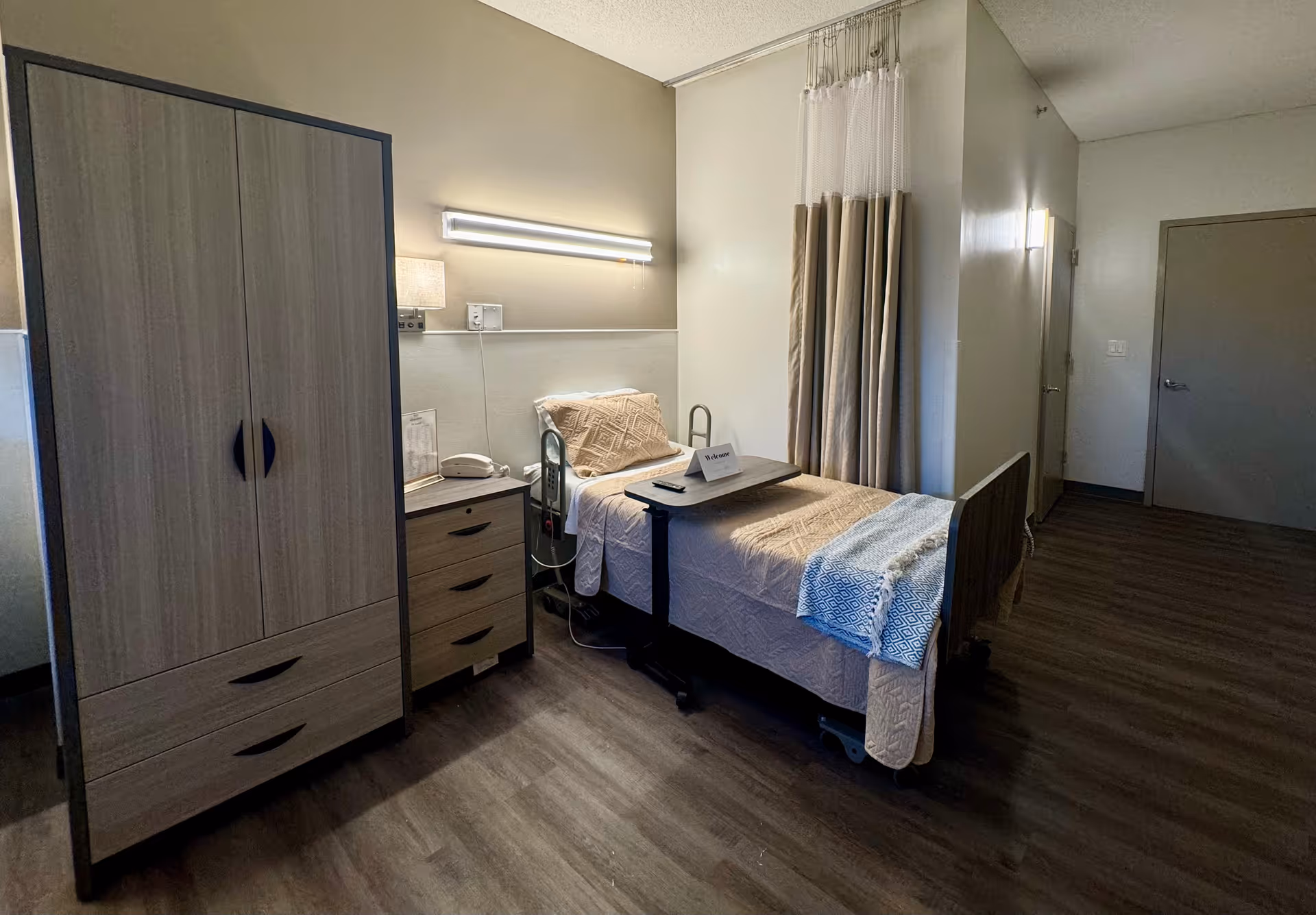 A simple resident bedroom with a single hospital-style bed, bedside table and wardrobe, and a partially drawn privacy curtain.
