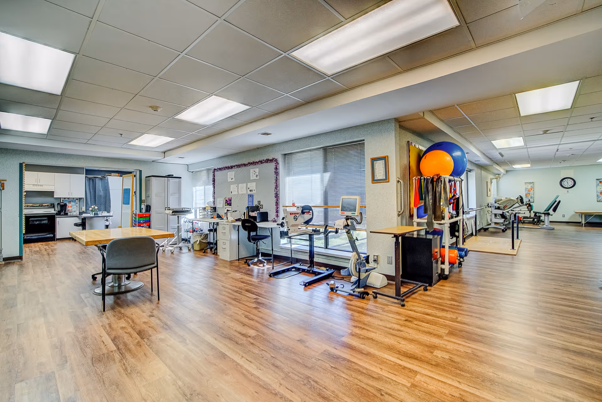 A spacious room with wooden flooring and fluorescent ceiling lights, featuring exercise equipment such as stationary bikes and therapy balls on racks. There is a table with chairs, a desk with office supplies, and a kitchenette area with a stove and refrigerator in the background. The room has large windows with blinds and appears to be a multi-purpose therapy or activity room.