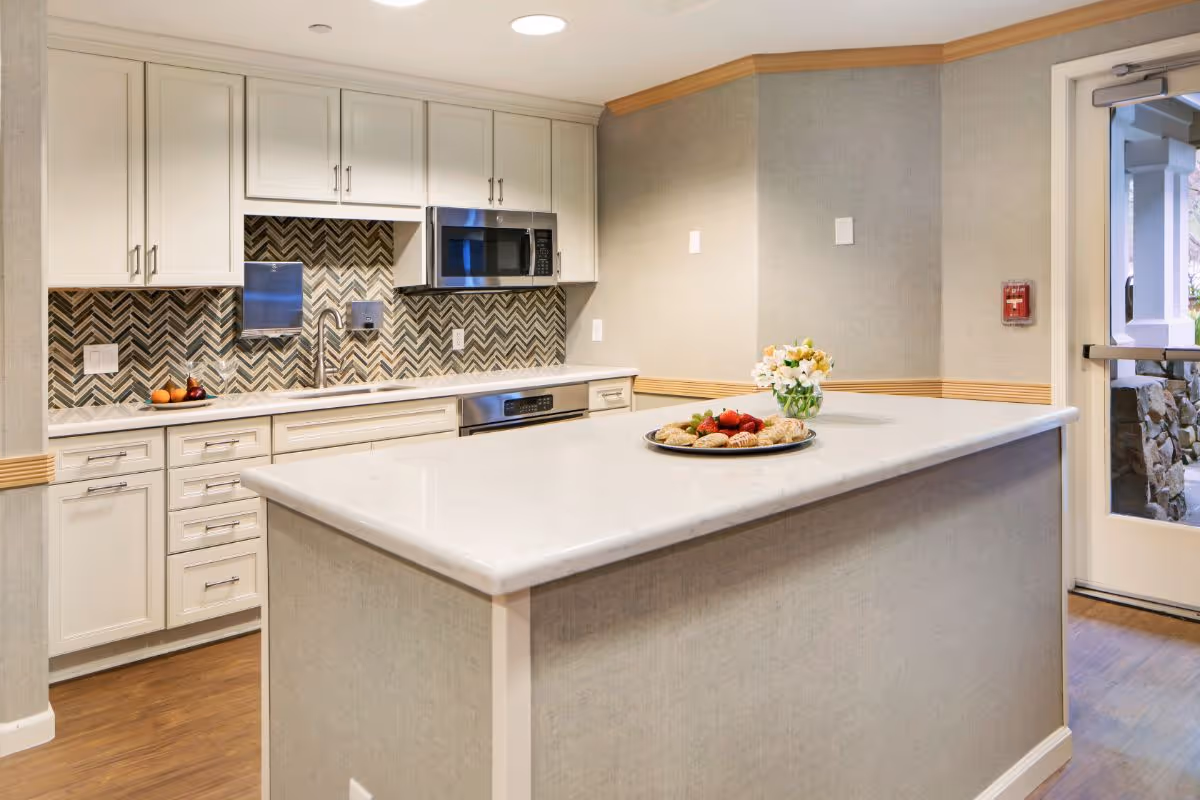 Modern kitchenette with a marble island topped with a platter of pastries, white cabinetry, and a chevron tile backsplash.