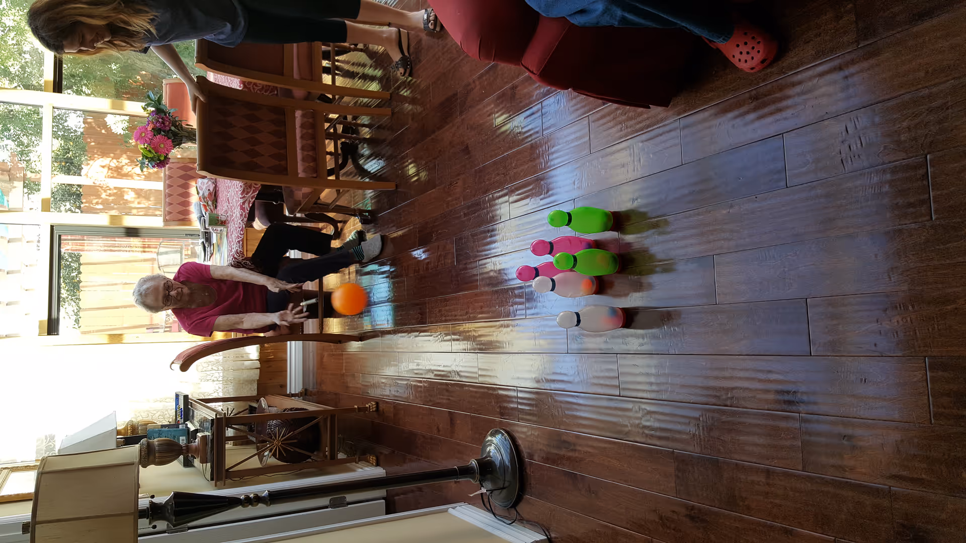 An elderly woman seated at a dining table rolls an orange ball towards a set of colorful plastic bowling pins arranged on a wooden floor. A younger woman stands nearby watching and smiling. The room has large windows with sheer curtains letting in natural light, a table with a floral tablecloth, and a floor lamp on the left side.