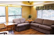 A cozy living room area with two patterned sofas arranged in an L-shape, a wooden side table with a decorative plant, large windows with blinds and valances allowing natural light to fill the room, and wooden flooring.