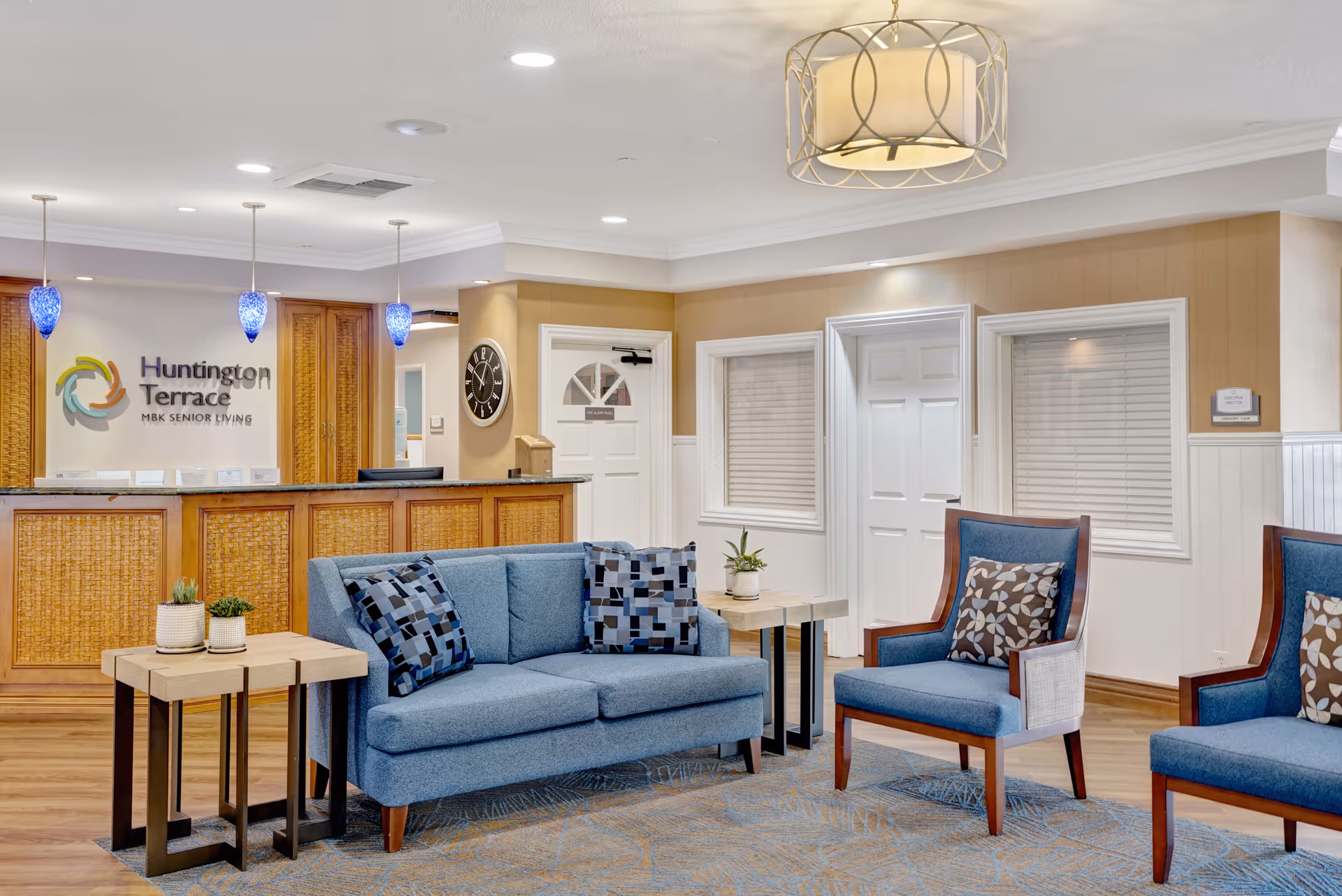 A bright and welcoming senior living facility lobby with a blue sofa and two matching armchairs arranged around two wooden side tables with small potted plants. The reception desk is in the background with a sign reading 'Huntington Terrace MBK Senior Living' on the wall behind it. The room features warm beige walls, recessed lighting, and a modern ceiling light fixture.