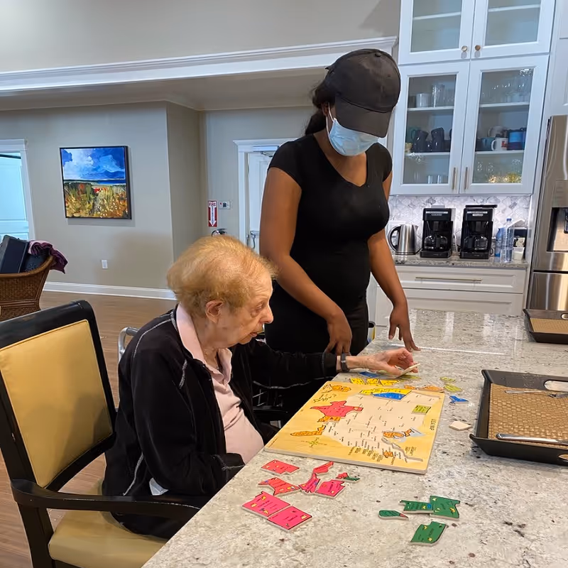 An elderly woman sitting at a kitchen island working on a colorful puzzle map, assisted by a standing caregiver wearing a black shirt, black cap, and face mask. The kitchen has white cabinets, a granite countertop, and coffee makers in the background.