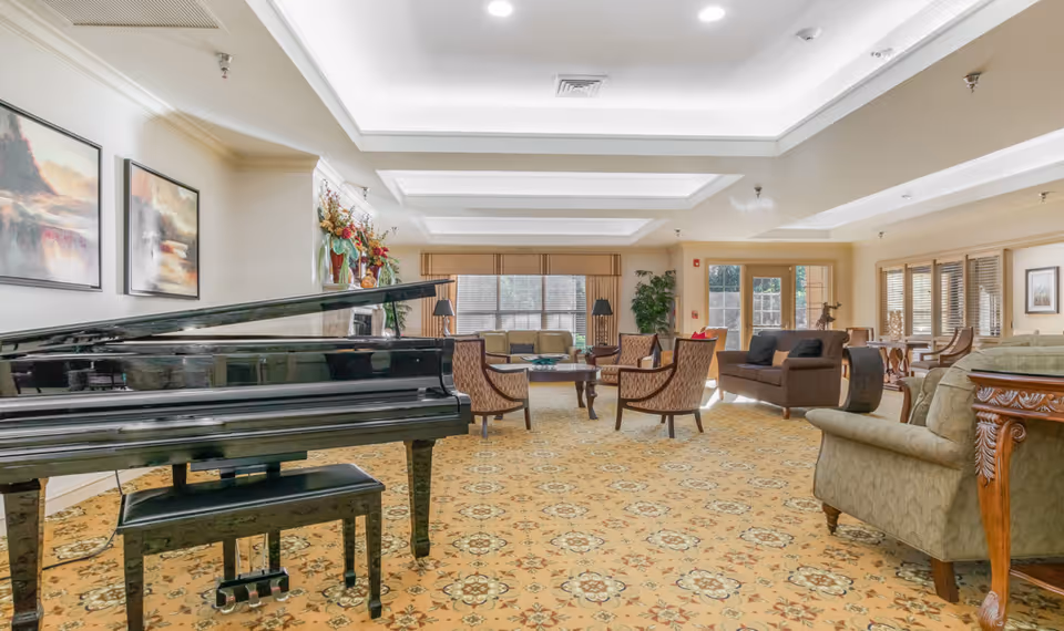 A spacious senior living community lounge featuring a black grand piano with a matching bench in the foreground. The room has patterned carpet flooring, several upholstered chairs and sofas arranged around coffee tables, large windows with blinds, and decorative plants and artwork on the walls. The ceiling has recessed lighting and a coffered design.