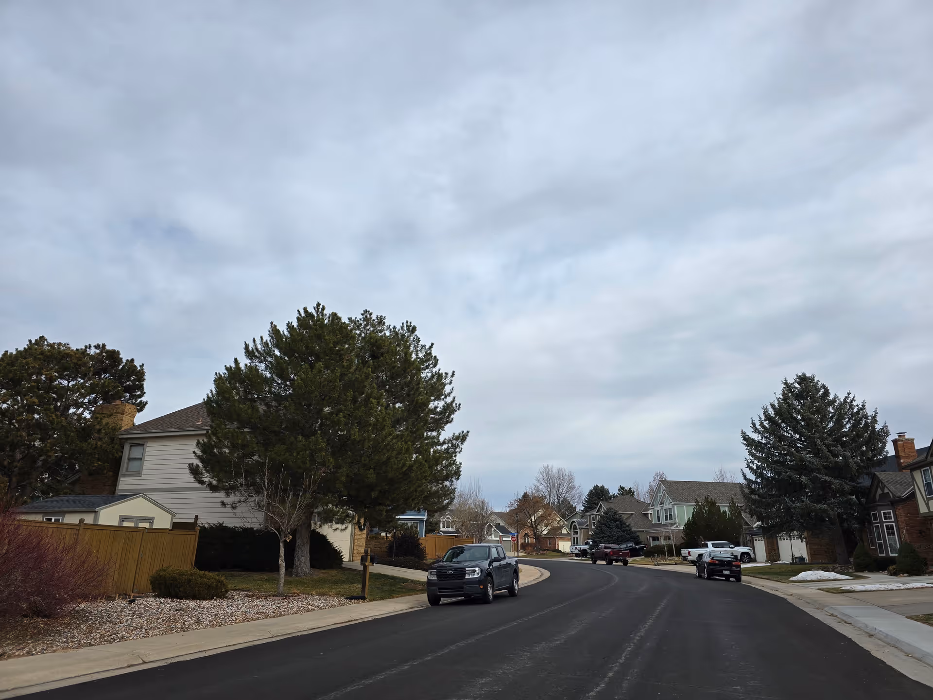 A suburban street with houses on both sides, several parked cars, trees, and a cloudy sky overhead.