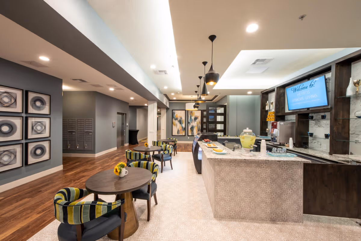 Interior view of a senior living facility lounge area with a refreshment counter on the right featuring a lemonade dispenser, cups, and snacks. Several round tables with striped upholstered chairs are arranged on the left side. The floor has a mix of wood and patterned tile. The walls are decorated with abstract art and a TV screen displaying a welcome message for Heartis Seniors Living.