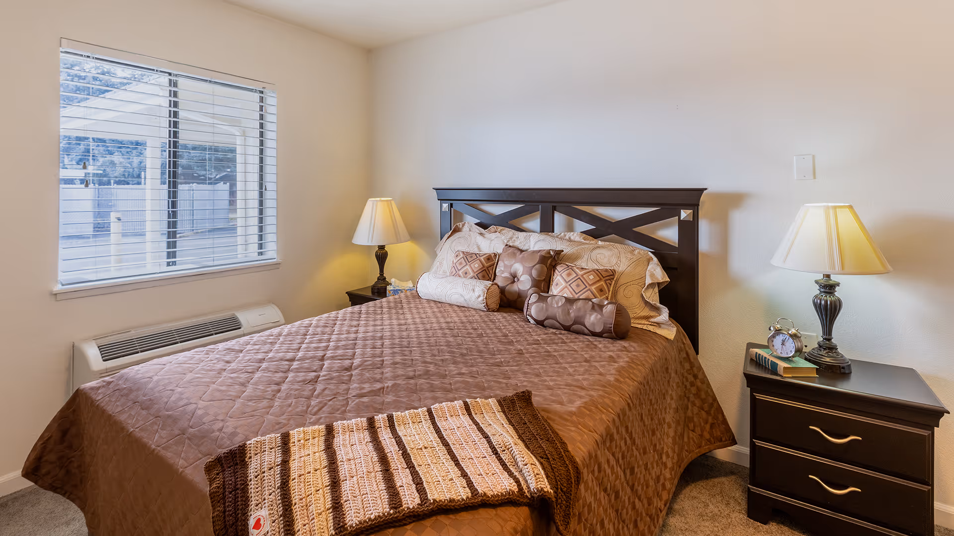 Sunlit bedroom with a double bed dressed in a brown quilt and decorative pillows, flanked by two nightstands and lamps.