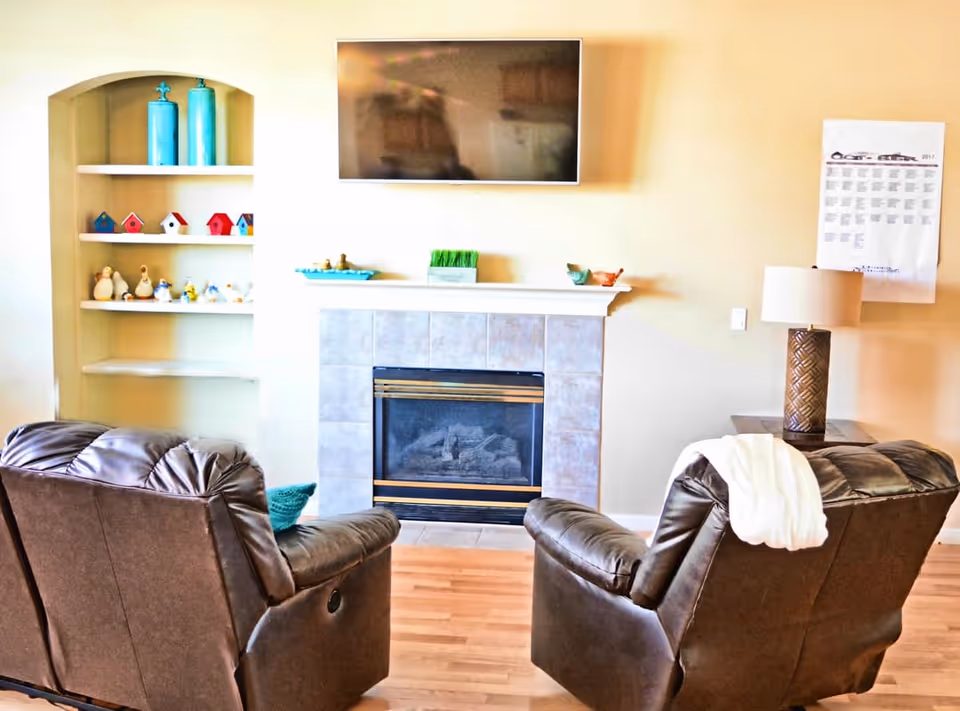 A cozy living room with two brown leather recliners facing a tiled fireplace. Above the fireplace is a mounted flat-screen TV. To the left of the fireplace is a built-in shelf with decorative items including small birdhouses and figurines. To the right is a side table with a lamp and a wall calendar. The floor is wooden.