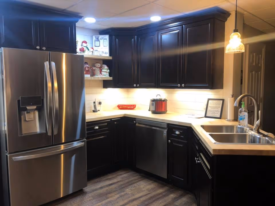 Modern kitchen with dark wooden cabinets, stainless steel refrigerator, dishwasher, and double sink. The countertop is light-colored, and there are decorative items on the shelves and counter. The kitchen is well-lit with ceiling lights and a hanging pendant light.