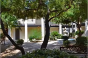 A shaded outdoor courtyard area with trees, bushes, and a bench. The background shows a building with white walls and a covered walkway supported by columns.