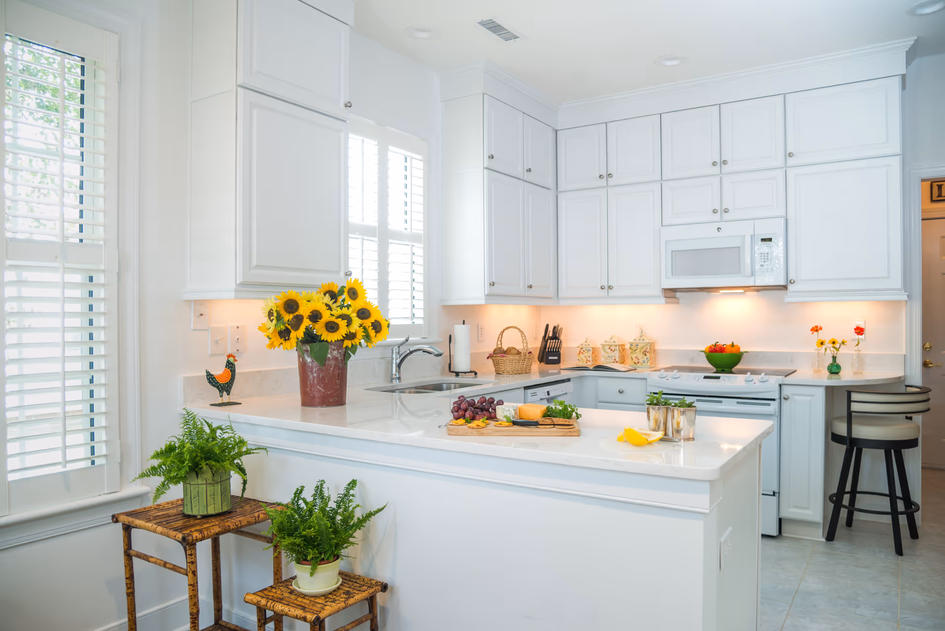 Bright and clean kitchen with white cabinets and countertops. A vase with sunflowers is placed on the counter near the window with white shutters. There are two small plants on wooden stools in front of the counter. The kitchen features a microwave, stove, and dishwasher, with a cutting board holding grapes, cheese, and herbs on the island. A couple of bar stools are positioned at the counter on the right side.
