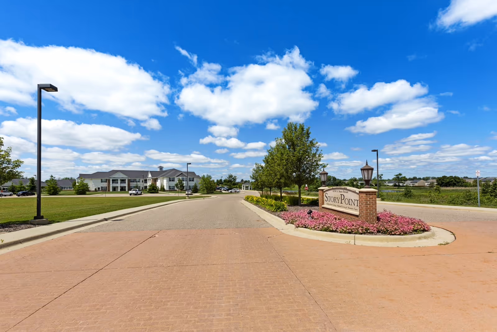 Entrance road leading to a large building with a manicured lawn and trees on a sunny day with blue sky and scattered clouds. A brick sign with flowers around it reads 'Story Point'.