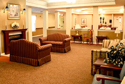 A cozy living room area in a senior living facility with patterned armchairs and sofas arranged around a fireplace. The room has warm lighting, beige walls, and carpeted flooring. There are small tables with lamps and framed artwork on the walls.