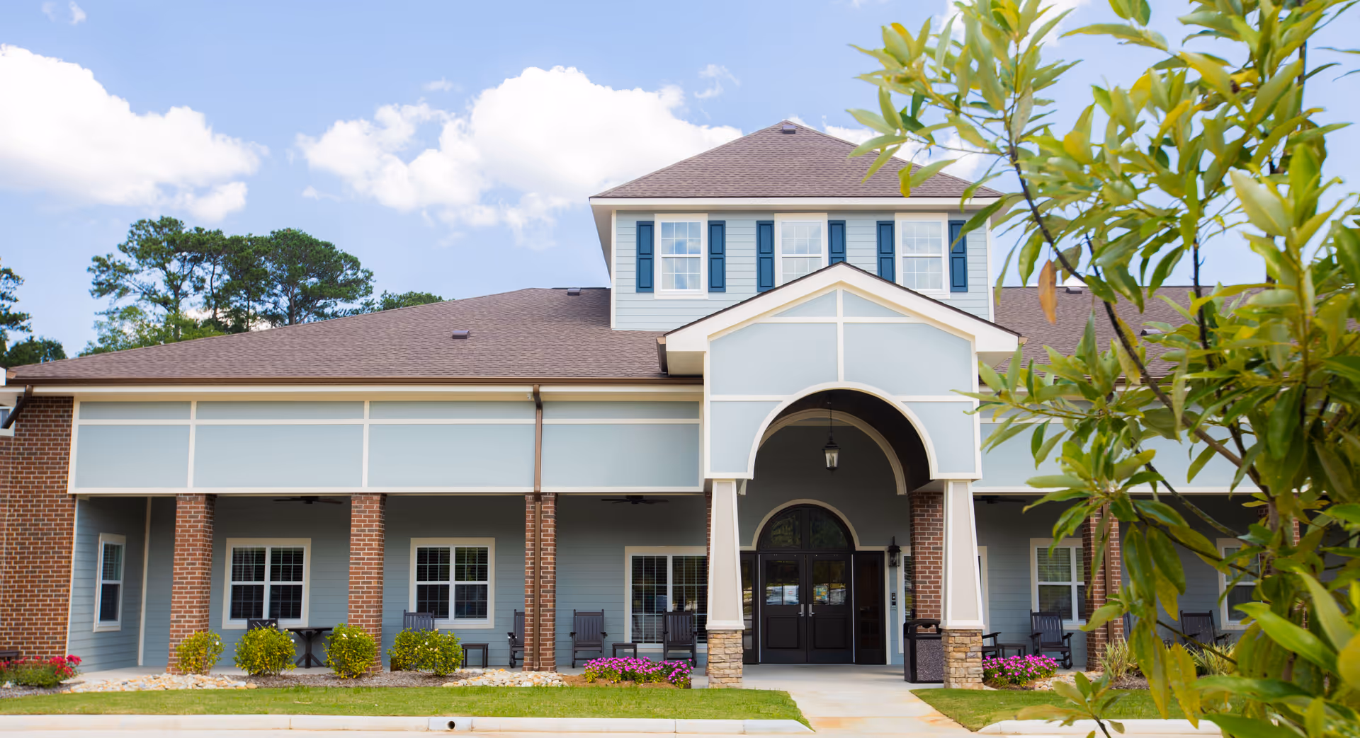 Front exterior view of a two-story senior living facility building with a covered entrance, brick pillars, blue siding, and several windows. There are plants and flowers in front of the building and a tree partially visible on the right side.