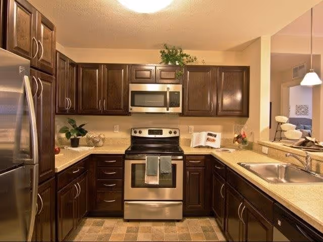 A modern kitchen with dark wooden cabinets, stainless steel appliances including a refrigerator, stove, and microwave. The countertops are light-colored with a sink on the right side. There are decorative plants and an open cookbook on the counter.