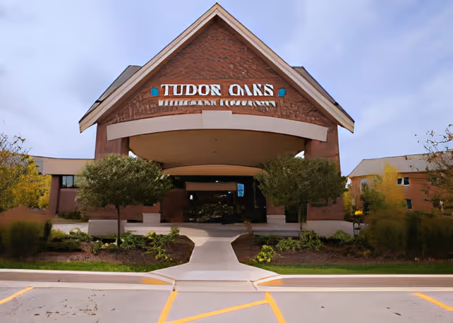 Entrance of the Tudor Oaks Senior Living Community building with a covered porte-cochère, signage, and landscaped walkway.