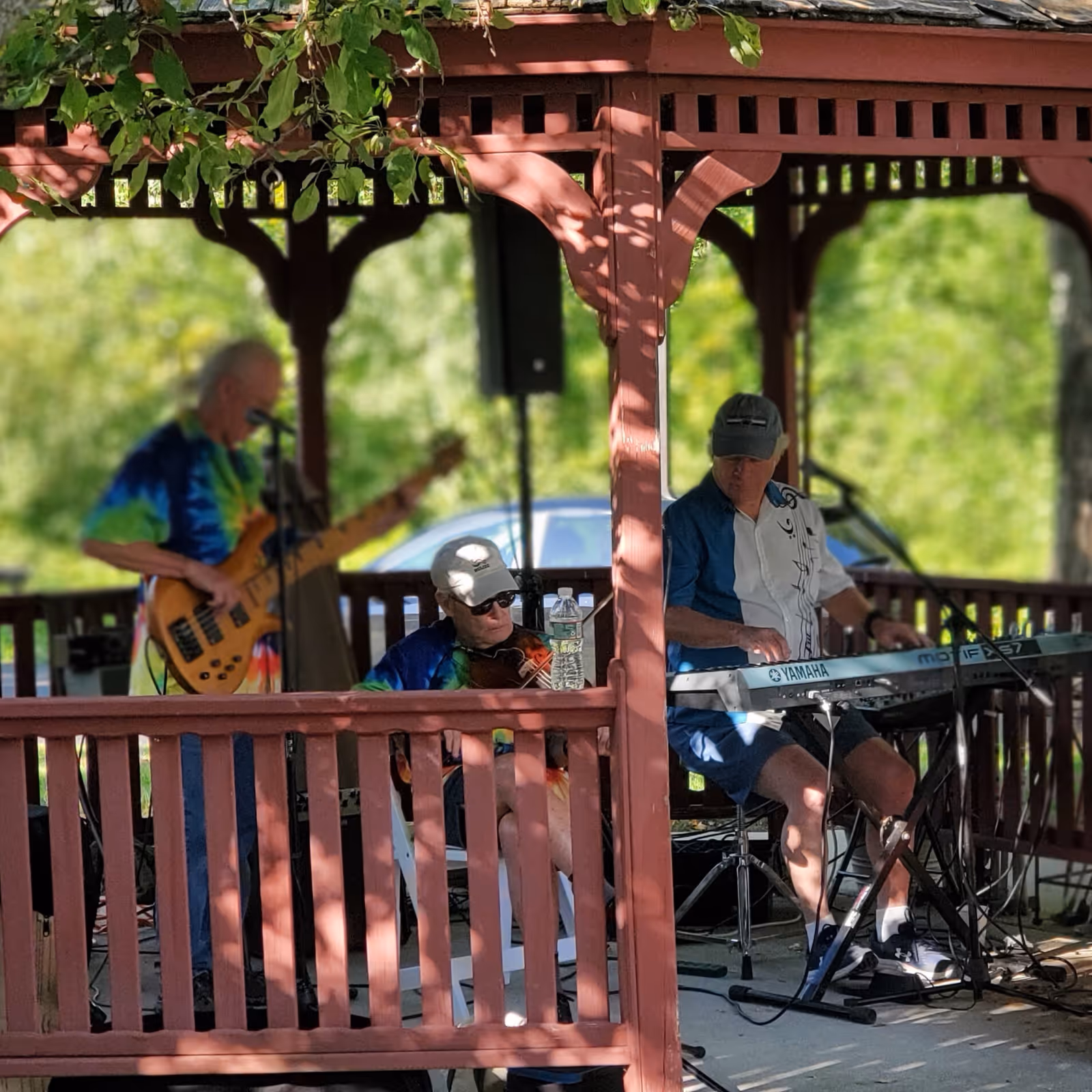Three elderly men playing musical instruments under a wooden gazebo outdoors. One man is playing a bass guitar, another is playing a violin, and the third is playing a keyboard. The setting is shaded with greenery in the background.
