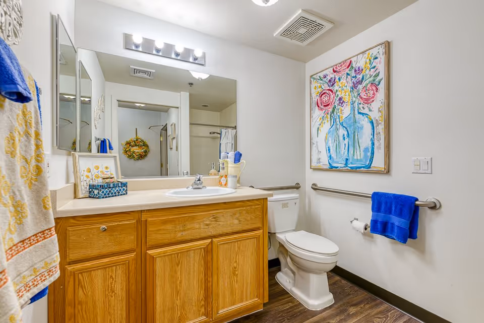 A clean bathroom with a wooden vanity cabinet, a sink, and a large mirror above it. There is a toilet next to the vanity with a blue towel hanging on a grab bar beside it. The walls are white, and there is a colorful painting of flowers in blue vases hanging above the toilet. The floor has a wood-like finish, and there are additional towels hanging on the left side.
