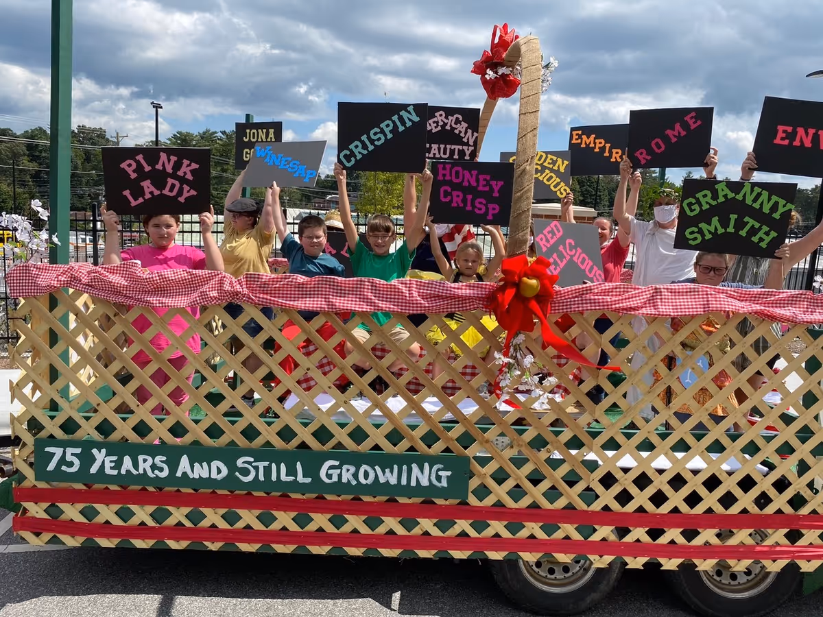 A group of children standing behind a wooden lattice fence decorated with red and white checkered fabric and a large red bow. Each child is holding a black sign with the name of an apple variety written in colorful letters, such as Pink Lady, Winesap, Crispin, Honey Crisp, Granny Smith, and others. A green sign on the fence reads '75 Years And Still Growing'. The scene is outdoors with a partly cloudy sky and trees in the background.