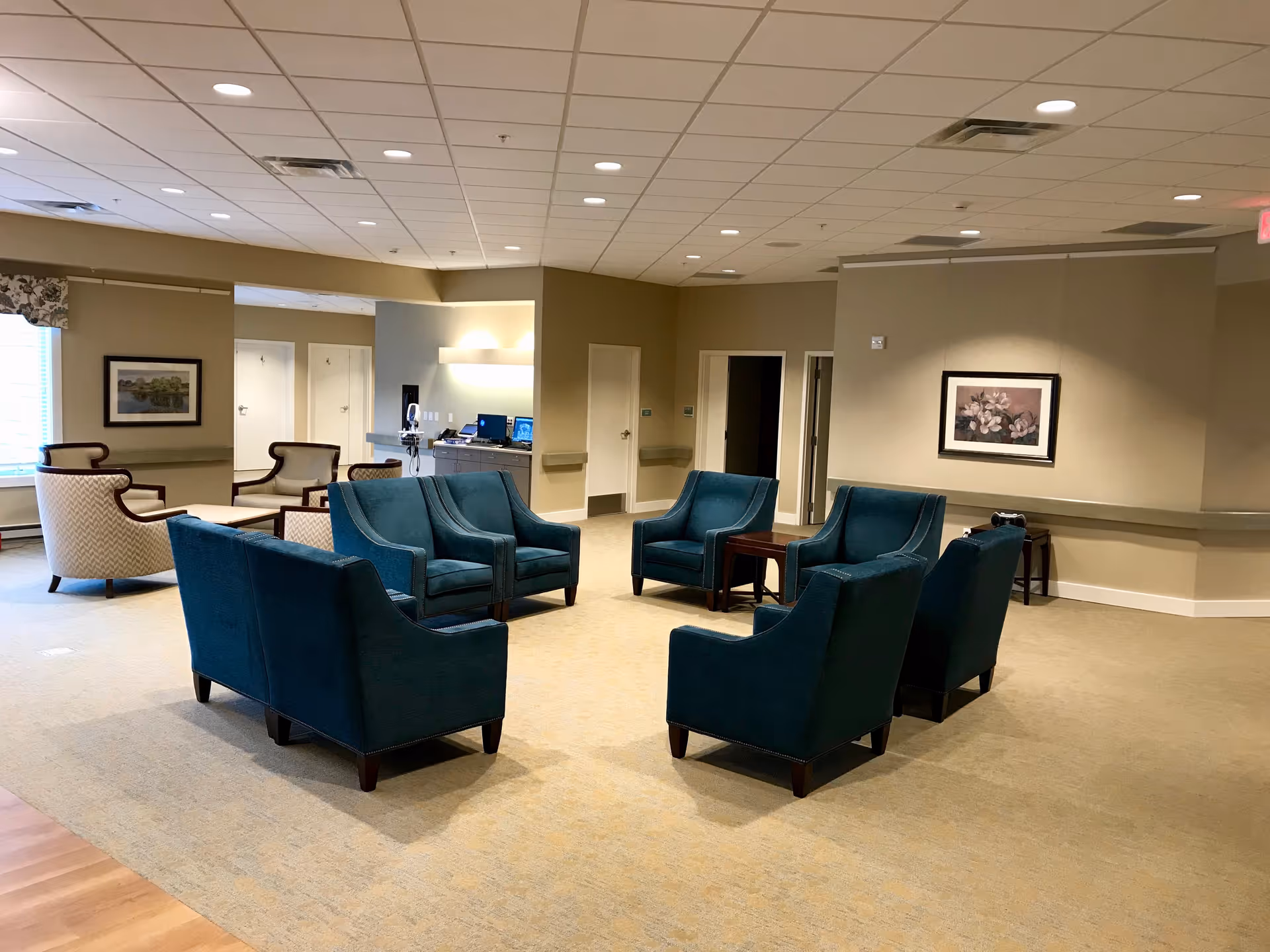 A spacious common area with several teal armchairs arranged in a circle around a small wooden table. In the background, there are additional beige armchairs near a window with patterned valances. The walls are beige with framed artwork, and the ceiling has recessed lighting. There is a small desk area with a computer and phone visible in the back.