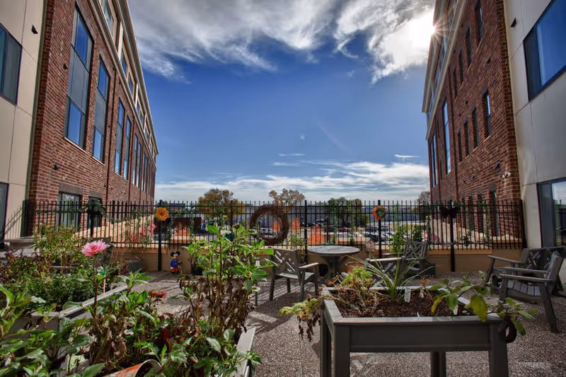 Outdoor courtyard patio with raised planters, seating and a table between two brick building wings under a partly cloudy sky.