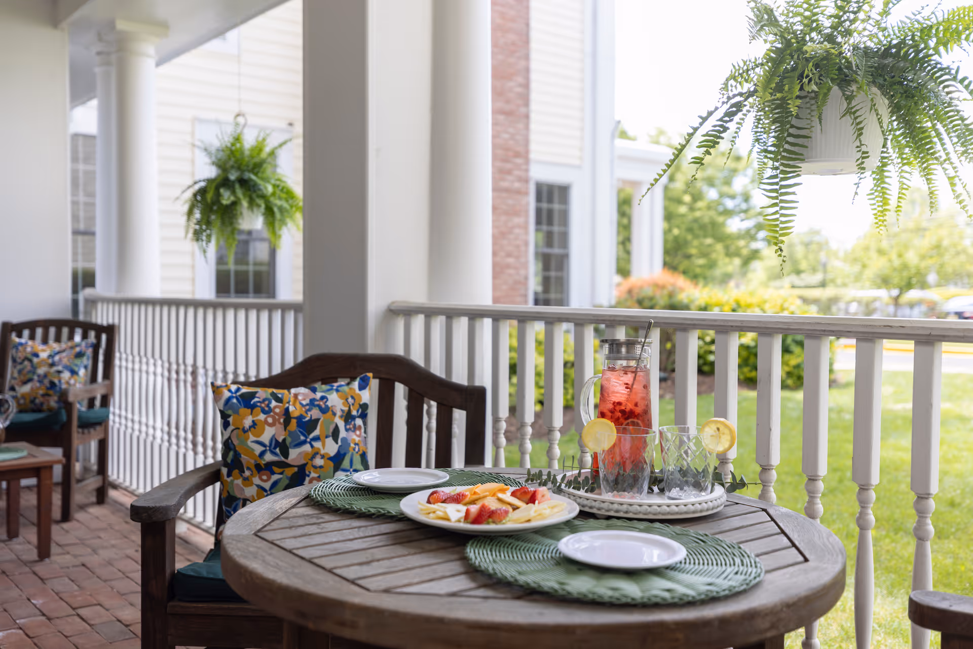 A cozy outdoor patio area with a round wooden table set with two green placemats, two white plates, a plate of sliced fruit, and a pitcher of red drink with lemon slices and two empty glasses. The table is surrounded by wooden chairs with colorful floral cushions. Hanging green plants are visible, and the background shows a white railing, brick pillars, and a green lawn with trees and shrubs.