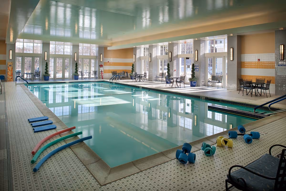 Indoor swimming pool area with clear water, pool noodles and foam dumbbells on the pool deck, several tables and chairs along the sides, large windows letting in natural light, and potted plants placed near the windows.