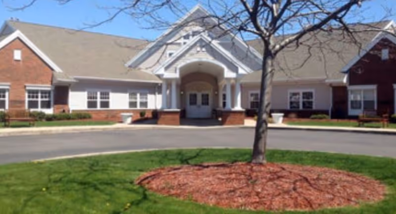 Front entrance of a single-story brick-and-siding senior living building with a covered portico, circular driveway, and a tree in a mulched island.