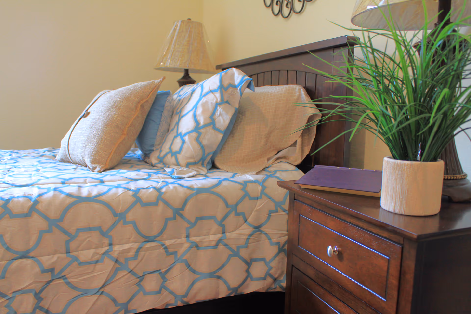 Bedroom in a senior living facility with a wooden headboard, blue patterned quilt, and plant.