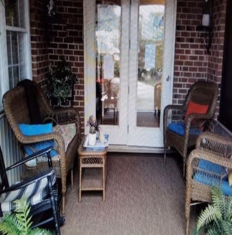 A covered outdoor patio area with four wicker chairs, each with colorful cushions, arranged around a small wicker table with a plant and decorative items. The patio has a brick wall and glass double doors leading inside. There are some plants visible in the foreground.